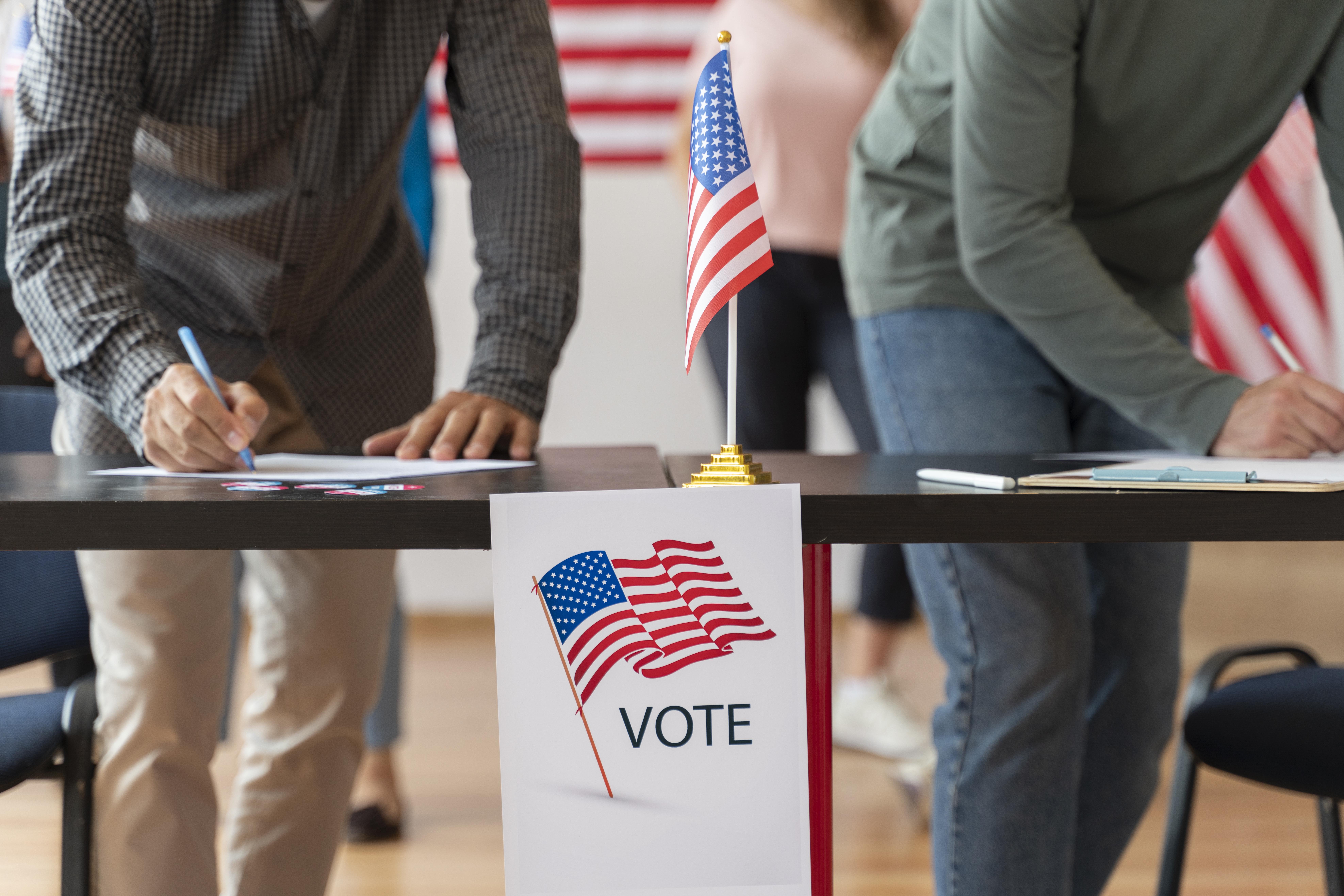 People filling out voting forms at a table with an American flag and a sign that says 'VOTE' in a polling station.