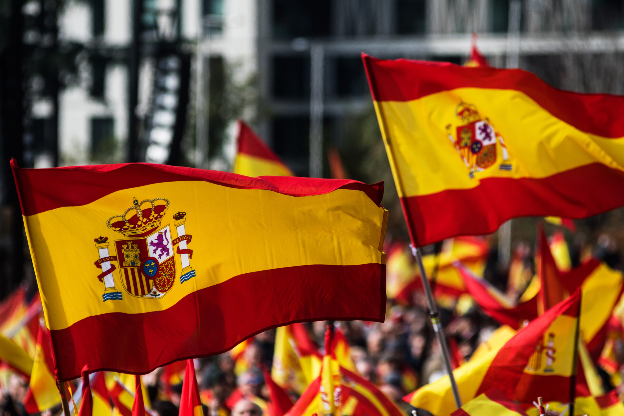 Spanish flags waving in a crowd, featuring the national coat of arms on a red and yellow background.