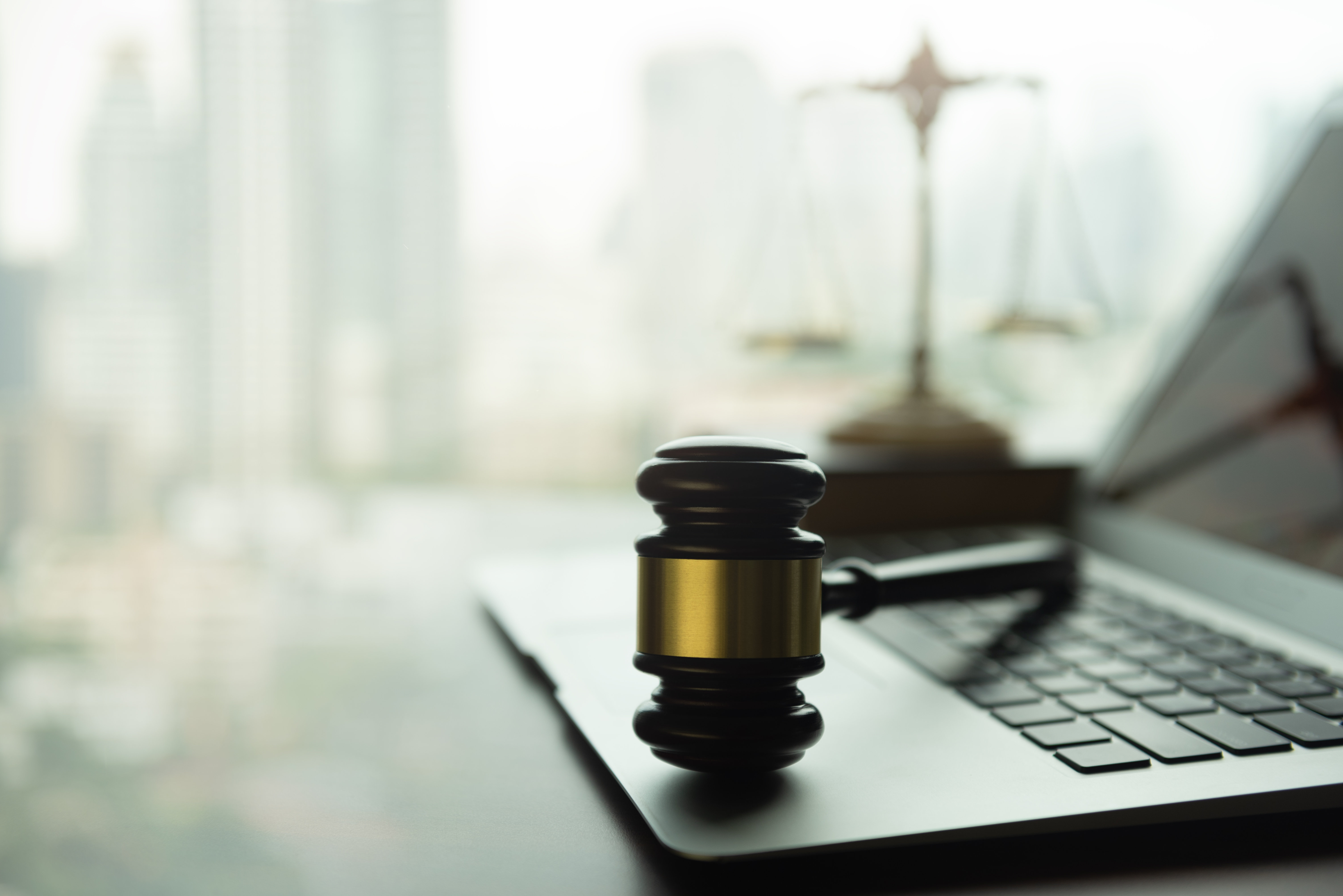 A gavel resting on a laptop keyboard with a blurred background showing a balance scale and a cityscape