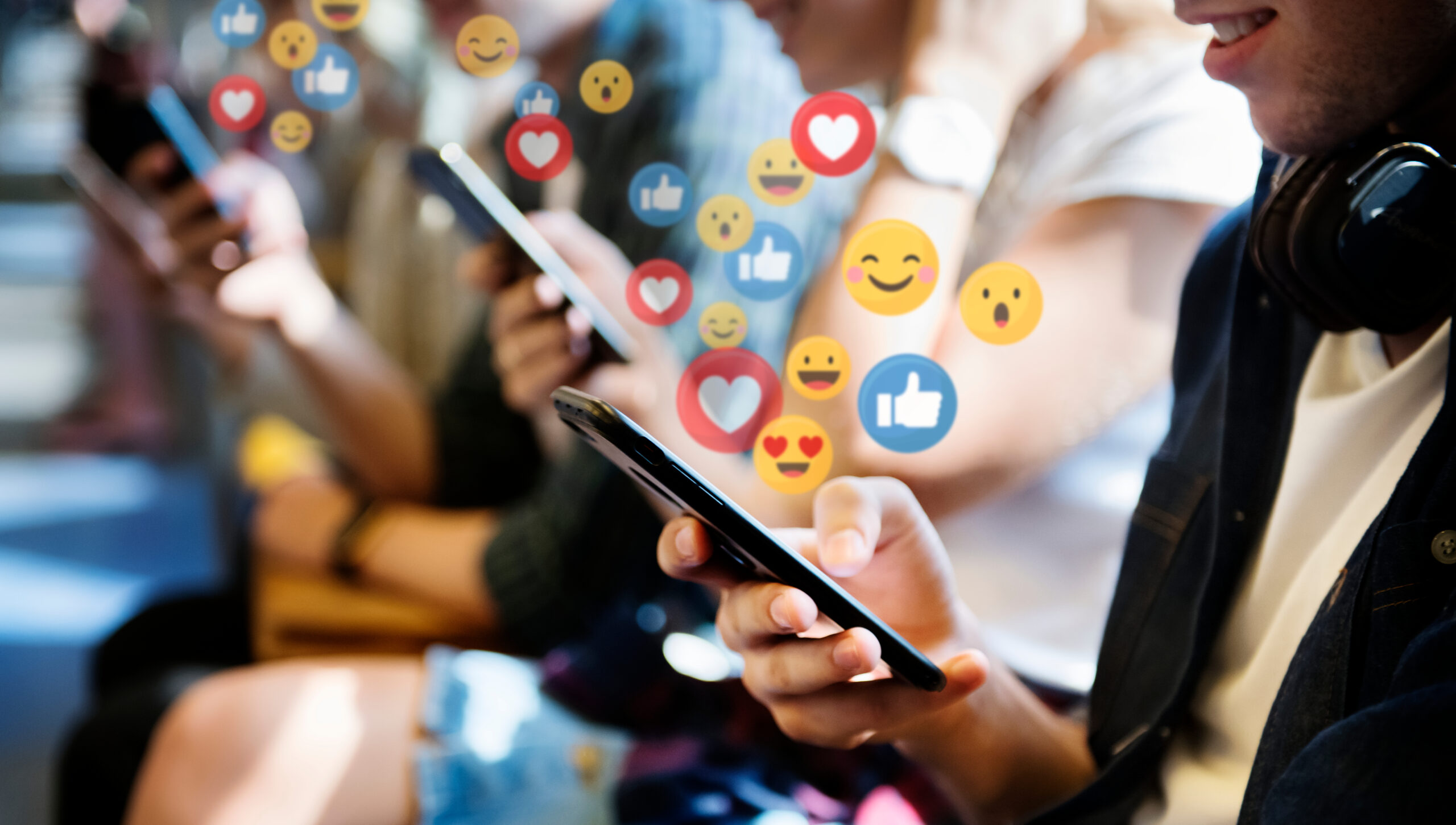 Group of people using smartphones with various social media reaction icons like hearts, likes, and emojis floating above.