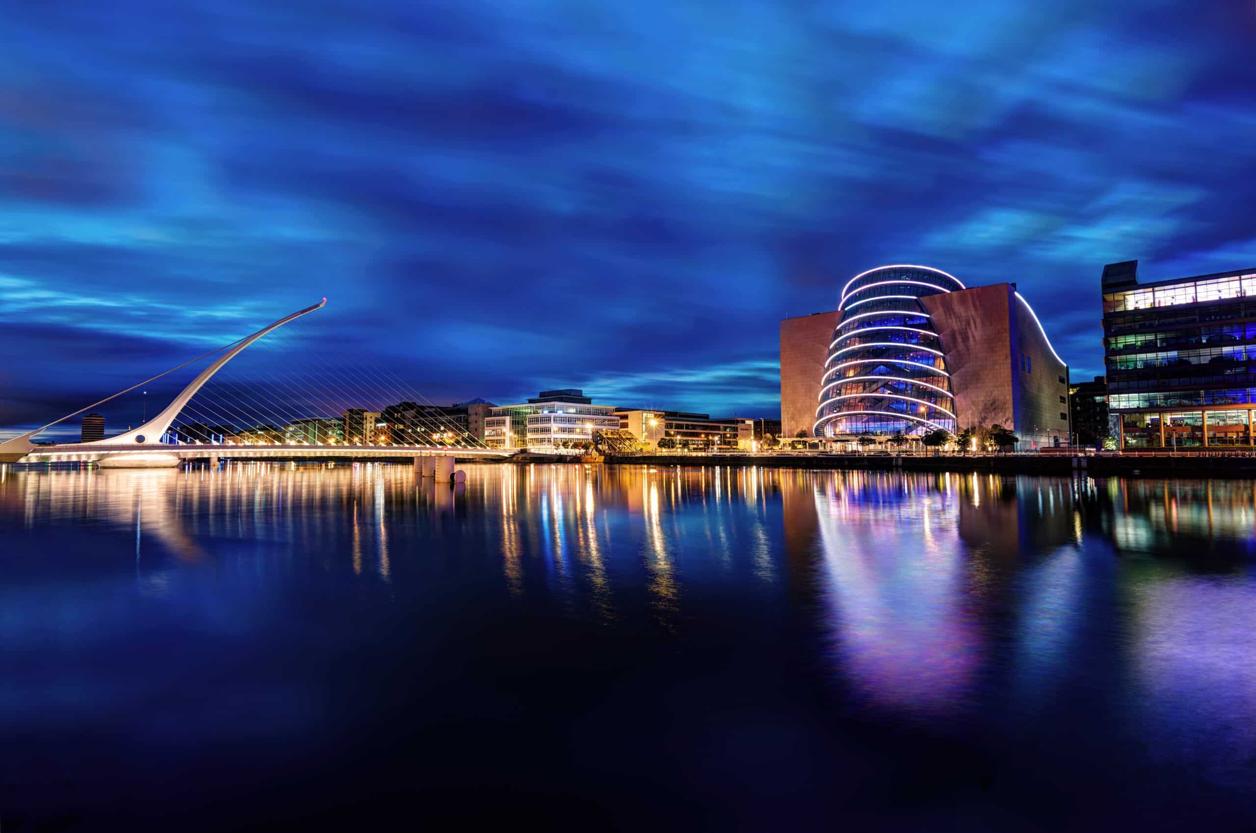 Dublin view of a modern cityscape with a brightly lit bridge and a distinctive, illuminated building reflected in the water.