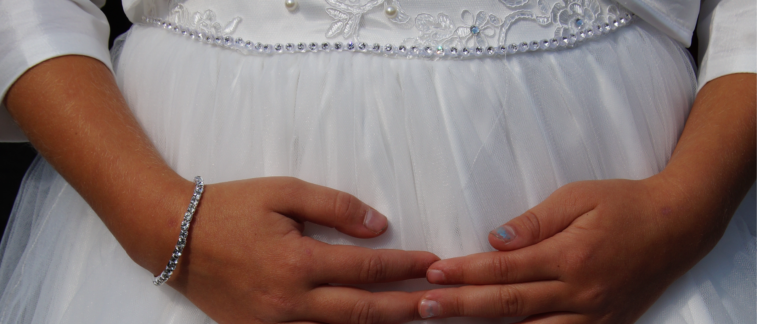 Close-up of a child bride's hands resting on her belly, wearing a white dress with pearl embellishments