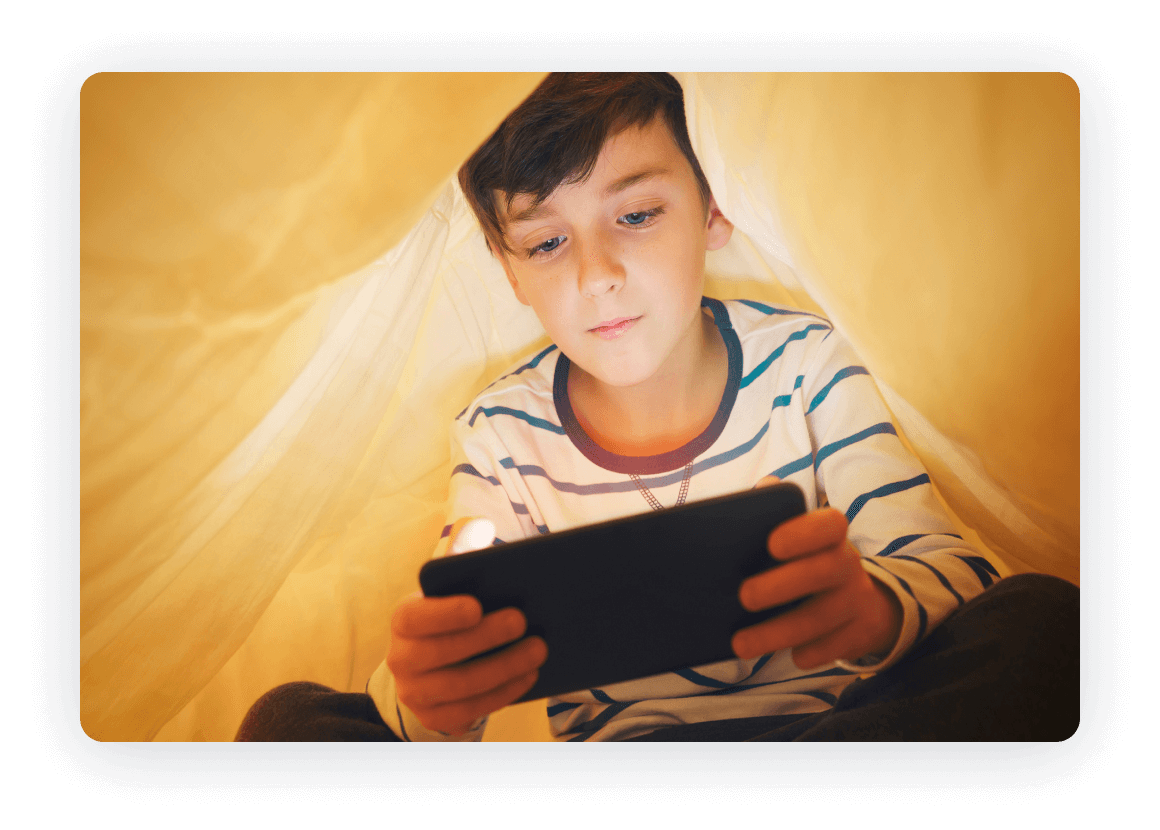 Young boy using a tablet while lying under a blanket.