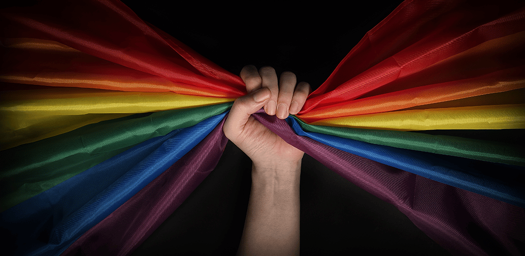 A hand gripping a pride flag with rainbow colors against a dark background, symbolizing inclusivity and support.